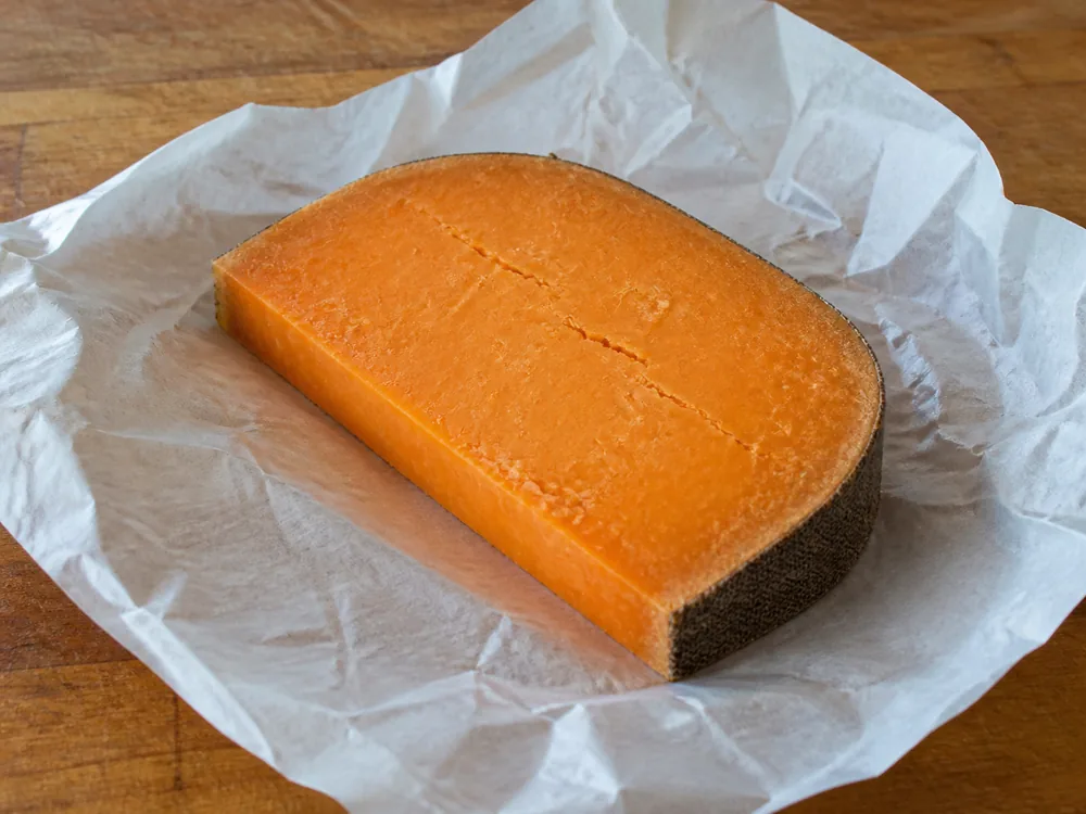 Cut piece on a wooden board loosely wrapped in waxed cheese paper with a dry cloth rind edge - Sparkenhoe Red Leicester | KnowTheCheese