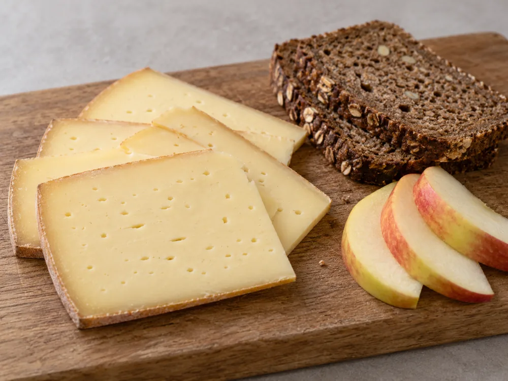 Slices of pale yellow cheese with small holes beside dark mixed rye bread and fruit slices - Tilsiter | KnowTheCheese