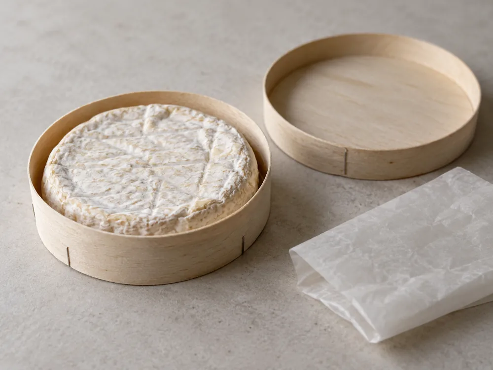 Illustration of a small round soft cheese with a wrinkled pale rind in a plain round wooden box, with the lid and a sheet of waxed paper beside it - Tunworth | KnowTheCheese
